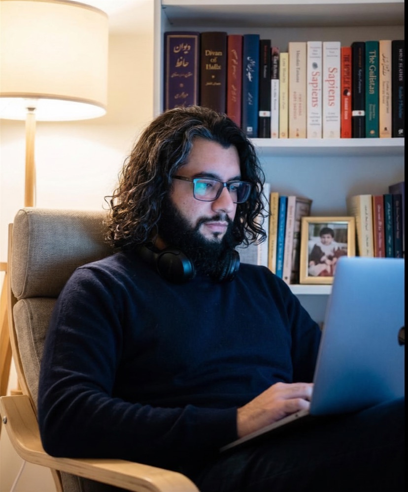 Saman in a home study, reading from a laptop, soft warm lamp light behind, a bookshelf in the background.